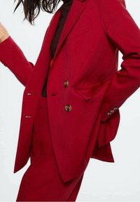 Femme portant un blazer rouge double boutonnage sur mesure et un pantalon rouge assorti sur un haut noir, posant avec une main sur la hanche.