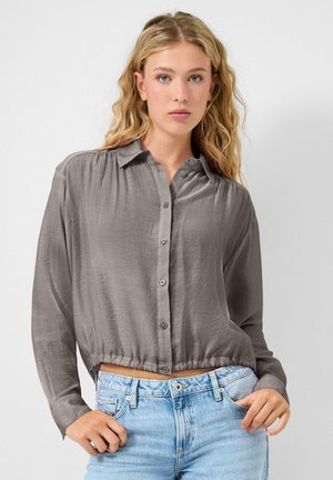 Jonge vrouw met golvend blond haar, gekleed in een loszittende grijze blouse met knopen en een lichtblauwe spijkerbroek, staand tegen een effen achtergrond.