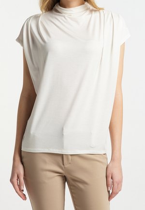 Woman wearing a white short-sleeve high-neck top and beige pants, standing with arms relaxed by her sides against a white background.