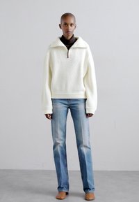 Pulover alb texturat cu guler cu fermoar, asortat cu blugi drepți de culoare bleu deschis. Culoare uniformă și croială relaxată.