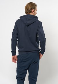 Mørkeblå hettegenser laget av bomullsblanding, med kengurulomme og ribbestrikkede mansjetter. Bakvinkelen viser et rent, minimalistisk design.