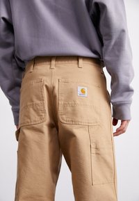Pantalons cargo beige avec plusieurs poches, fabriqués dans un tissu durable. L'arrière présente une étiquette carrée avec un logo et des détails de couture.