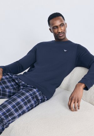 Homme en sweat-shirt bleu marine et pantalon à carreaux bleu s'inclinant sur un canapé clair à texture contre un fond uni.