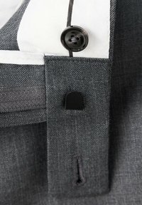 Gros plan d’un bouton noir sur un tissu gris avec une boutonnière et une partie d’une chemise blanche et noire rayée en dessous.