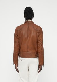 Καφέ δερμάτινο μπουφάν μηχανής με ραμμένες λεπτομέρειες στους ώμους και τους αγκώνες, εφαρμοστό σιλουέτα και διακοσμητικά αγκράφες στα πλαϊνά, εμφανιζόμενο από την πίσω πλευρά.