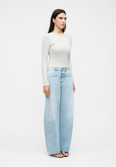 Γυναίκα με μακριά σκουρόχρωμα μαλλιά φοράει ανοιχτογκρι μακρυμάνικο τοπ και φαρδιά ανοιχτά μπλε τζιν, στέκεται ξυπόλητη σε απλό λευκό φόντο.