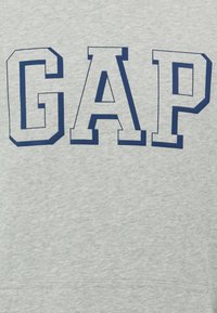 Sudadera gris con un audaz logo "GAP" en azul marino con fuente deportiva y en bloque. El material tiene una textura suave, con un dobladillo acanalado para mayor detalle.
