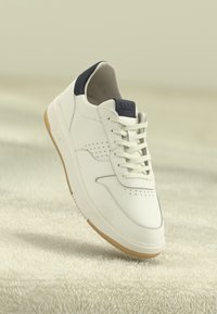 White low-top sneaker with beige sole, black heel tab, and laces, suspended above textured beige surface.