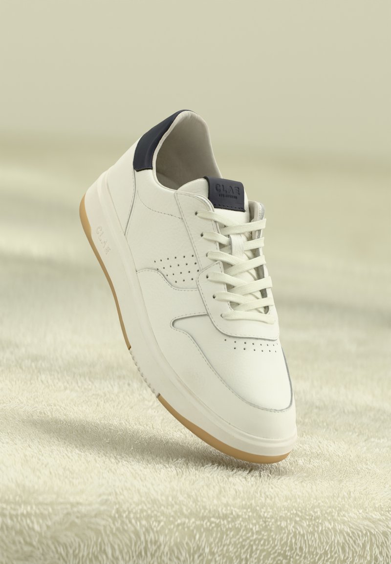 White low-top sneaker with beige sole, black heel tab, and laces, suspended above textured beige surface.