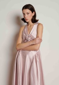 Γυναίκα με αμάνικο λαμπερό ανοιχτό ροζ φόρεμα με λουλουδάτη λεπτομέρεια στο στήθος, με σταυρωμένα χέρια, στέκεται μπροστά σε απαλό ανοιχτόχρωμο τοίχο.