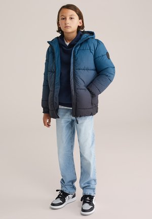 Veste matelassée en bleu et noir, dotée d'une capuche, d'une fermeture éclair et de poignets élastiques. Portée par-dessus un sweat à capuche bleu marine, avec un jean clair et des baskets.