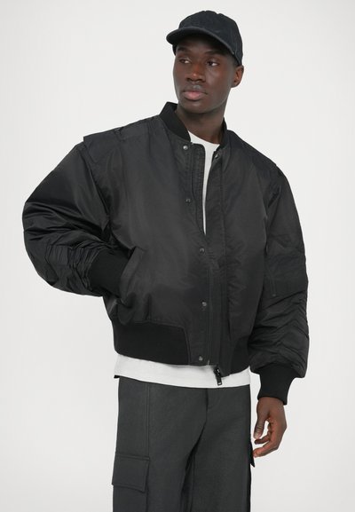 Juodas bombonešio striukė, pasižyminti blizgiu, lygiu paviršiumi, ribbintomis rankogaliais ir apačia bei užtrauktuku priekyje su spaustukų akcentais.