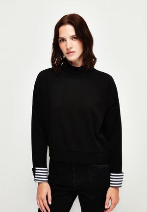 Pullover nero con collo alto, spalle scese e polsini a righe a contrasto. Tessuto morbido con una vestibilità comoda e cuciture minime.
