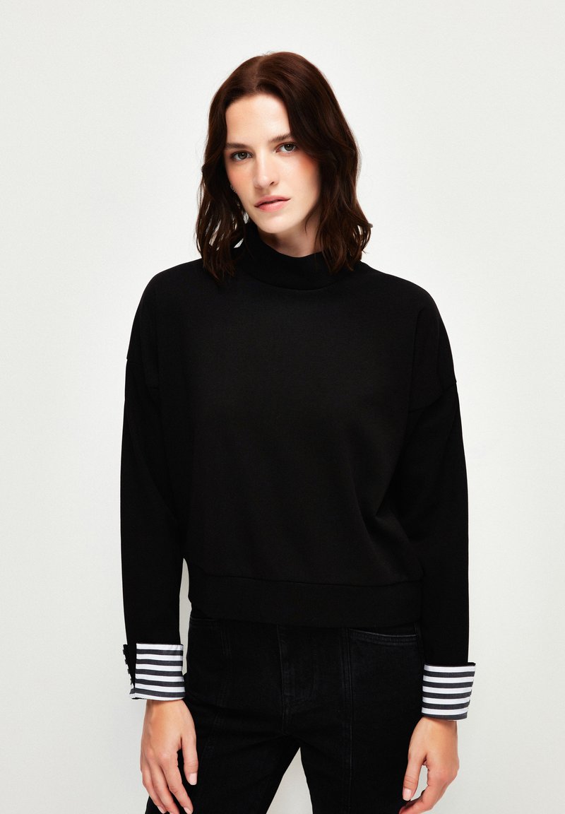 Schwarzer Sweatshirt mit hohem Kragen, fallenden Schultern und kontrastierenden gestreiften Bündchen. Weicher Stoff mit lockerer Passform und minimalen Nähten.