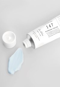 Barrierekremtube i hvitt med sølv dyse. Lys blå krem renner ut av åpningen. Lokket er på en hvit bakgrunn.