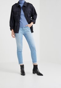 Chaqueta de utilidad negra con grandes bolsillos, cuello de tortuga azul acanalado, vaqueros de mezclilla azul claro y botas negras hasta el tobillo con tacón cuadrado.