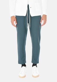 Uomo che indossa pantaloni teal con coulisse, un cardigan bianco a maglia, calzini neri e sneakers bianche, in piedi davanti a uno sfondo neutro.