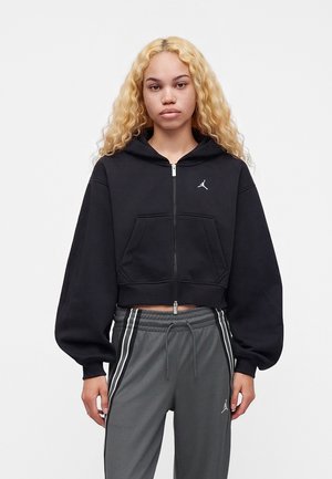 Jonge vrouw met lang, krullend blond haar, die een zwarte cropped zip hoodie en grijze broek met trekkoord en witte strepen draagt, staand tegen een effen achtergrond.