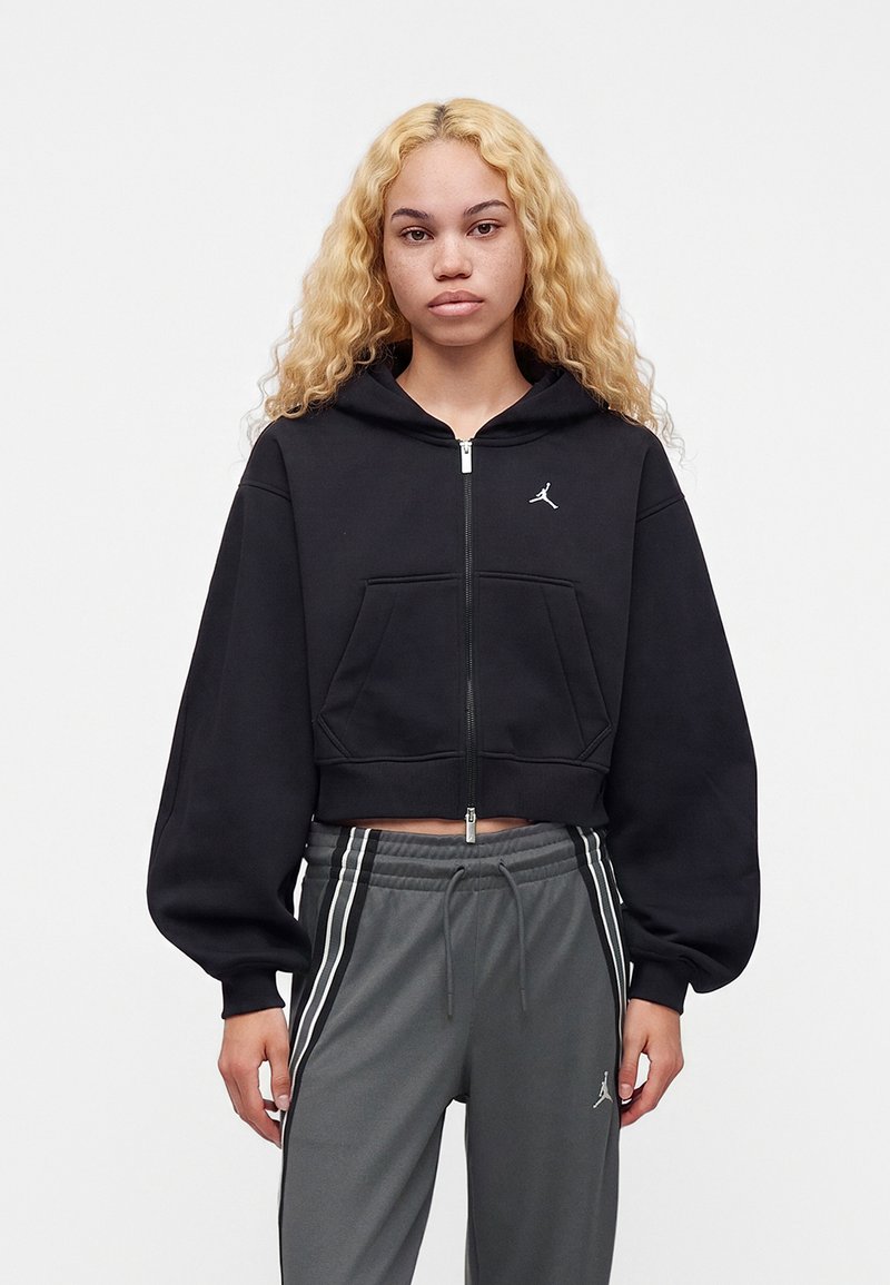 Jeune femme aux longs cheveux blonds bouclés portant un sweat à capuche court noir zippé et un pantalon gris à cordon avec des bandes blanches, debout devant un fond uni.