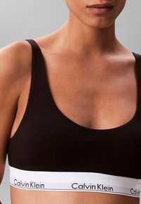 Zbliżenie osoby noszącej czarny sportowy biustonosz Calvin Klein z białą elastyczną taśmą z nazwą marki.