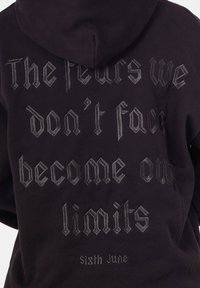 Zwarte hoodie met capuchon, met zilverkleurige gotische tekst op de achterkant: "De angsten die we niet onder ogen zien, worden onze grenzen. Sixth June." Zachte textuur.