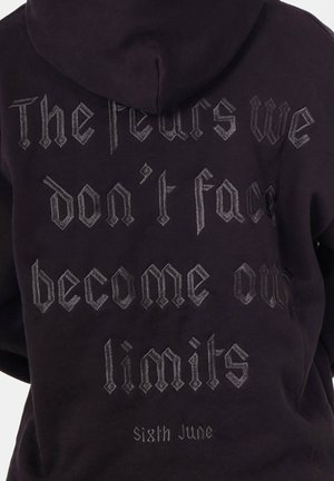 Zwarte hoodie met capuchon, met zilverkleurige gotische tekst op de achterkant: "De angsten die we niet onder ogen zien, worden onze grenzen. Sixth June." Zachte textuur.