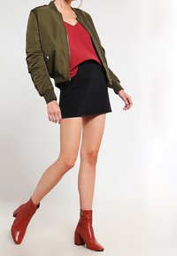 Olivově zelená bomber bunda, červený top s výstřihem do v, černá mini sukně a červené boty na kotníky s blokovým podpatkem. Hladké textury a ležérní styl.