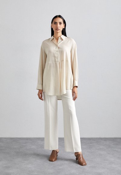 Camicia button-up beige chiaro con vestibilità rilassata, maniche lunghe e orlo arrotondato, abbinata a pantaloni bianchi a gamba larga e scarpe slip-on marroni.