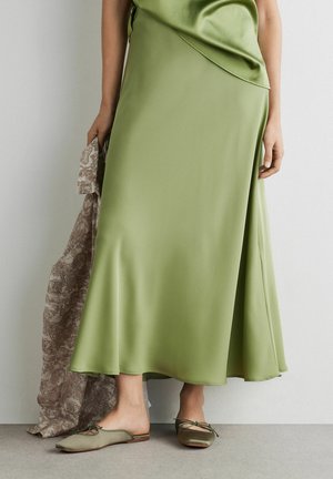 Kvinde iført en lang, lysegrøn satin nederdel og matchende top, der holder et mønstret brunt stof og står barfodet på en grå gulv.