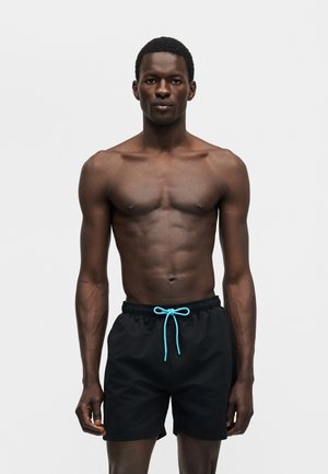 Jeune homme athlétique debout, portant un short de bain noir avec un cordon bleu vif, sur un fond gris clair uni.