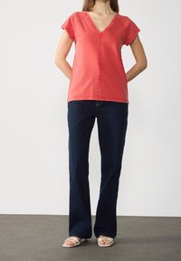 Rød bomulds V-hals top med korte ærmer, parret med mørkeblå flare jeans og hvide sandaler med stropper, mod en ensfarvet baggrund.
