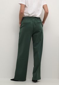 Pantalons larges verts avec une taille élastique, texture lisse et poches arrière. Associés à des chaussures noires à bout pointu.