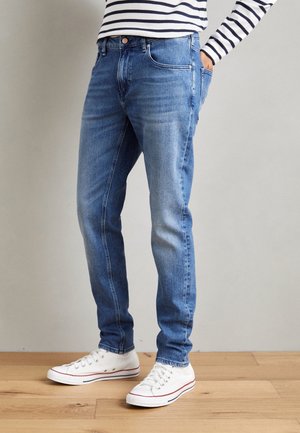 Persoon die blauwe slim-fit jeans, witte sneakers en een wit overhemd met lange mouwen met zwarte horizontale strepen draagt, staand op een houten vloer.