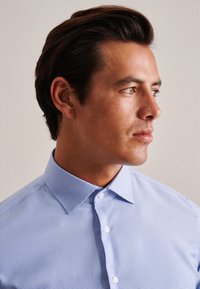 Jasnoniebieska koszula w kratkę z klasycznym kołnierzykiem, zapinana na guziki, o fakturowanym materiale, prezentowana na męskim modelu patrzącym w bok.