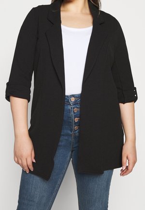 Vrouw draagt een zwarte blazer met open voorkant en opgestroopte mouwen over een wit topje en een hoge taille blauwe spijkerbroek met vier zichtbare knopen.