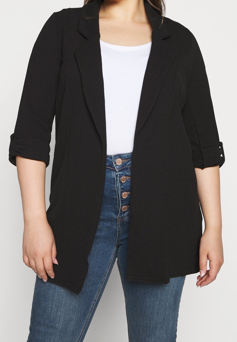 Femme portant un blazer noir ouvert à l’avant avec manches retroussées sur un top blanc et un jean bleu taille haute avec quatre boutons visibles.