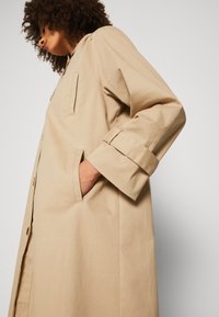 Palton din bumbac bej, cu croi drept, cu unul sau două rânduri de nasturi, buzunare pe laterale și mâneci răsucite. Textură netedă și design minimalist.