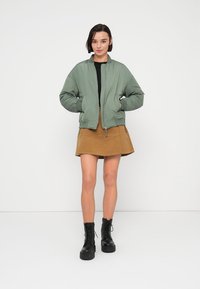 Πράσινο μπουφάν bomber με κλείσιμο με φερμουάρ, φορεμένο πάνω από μαύρο τοπ και φούστα σε σιλουέτα Α χρώματος ταμπά. Συνδυασμένο με μαύρα αρβυλάκια με κορδόνια.