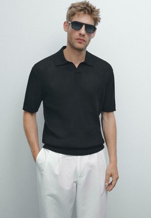 Polo shirt nero a righe con maniche corte e colletto classico, abbinato a pantaloni bianchi con pieghe. Il modello indossa occhiali da sole scuri.