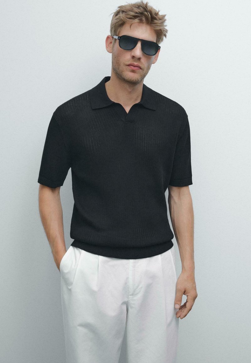 Černé žebrované polo tričko s krátkým rukávem a klasickým límcem, doplněné o bílé skládací kalhoty. Model má tmavé sluneční brýle.