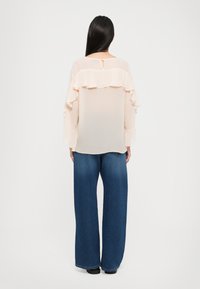 Lång, genomskinlig ljusrosa blus med volangdetaljer på baksidan, parat med vida blå jeans. Outfitten har en NYCKELHÅLSdetalj vid halsen.