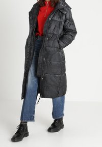 Dlouhý černý péřový kabát s kapucí, prošívanou strukturou, knoflíky vpředu a bočními kapsami, doplněný červeným topem a modrými džínami, doplněný černými botami.