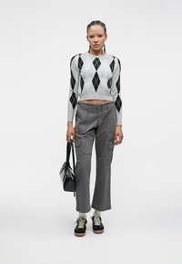 Femme avec des cheveux tressés portant un pull cropped en argyle, un pantalon cargo gris, des baskets noires et tenant un sac à main noir.