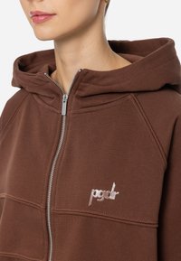 Ruda zip-up hoodie su didele kauke, su briaunomis detalėmis ir įsiūtu šviesiu logotipu. Audinys atrodo minkštas ir tekstūruotas.