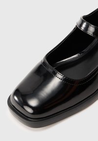 Zapato Mary Jane de charol negro con punta redonda, suela plana y doble tira. Acabado suave con sutiles detalles de costura.