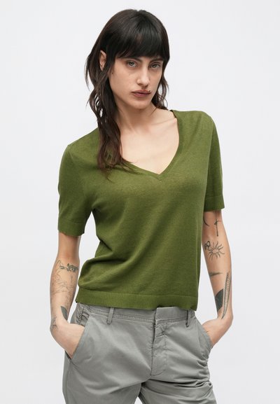 Πράσινο πουλόβερ με κοντά μανίκια και V-λαιμόκοψη, κατασκευασμένο από μαλακό υλικό. Το μοντέλο φοράει γκρι παντελόνι, αποκαλύπτοντας περίπλοκα τατουάζ στα μπράτσα.
