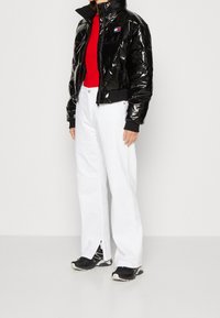 Giacca corta trapuntata nera lucida con colletto alto, con un piccolo logo sul petto, abbinata a pantaloni bianchi a gamba larga e sneakers nere.