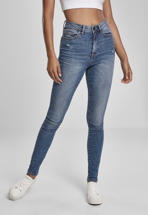 Femme portant un jean skinny bleu taille haute légèrement usé, des baskets blanches et un top court blanc, debout sur un fond gris.