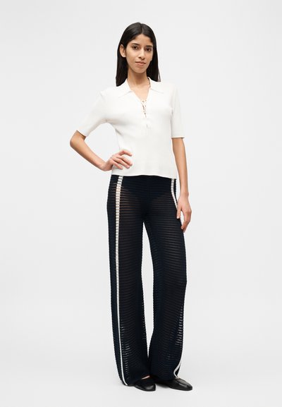 Λευκό κοντομάνικο ριμπ τοπ με γιακά και λεπτομέρεια δέσιμου, σε συνδυασμό με μαύρο διάφανο παντελόνι με φαρδιά πόδια και λευκές πλαϊνές ρίγες.
