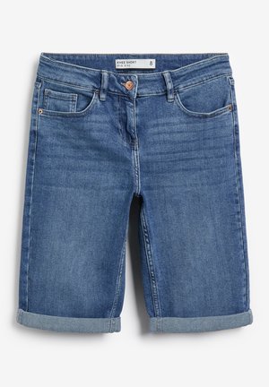 Vue arrière d'une personne portant un short en denim bleu de longueur moyenne avec des revers roulés et un haut blanc, main reposant sur le côté.